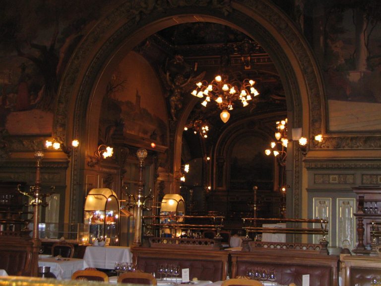 Ornate restaurant interior with arched doorways, golden decorations, and classical paintings evoke the elegance of France. Warm lighting from chandeliers reflects on mirrored surfaces and set tables, creating a luxurious, historic ambiance for any first time visitor.