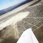 Aerial view of a large desert encampment at BurningMan, arranged in a semi-circular pattern with rows of tents and vehicles. The arid, sandy landscape stretches to distant mountains. Part of an airplane wing is visible in these unique Burning Man photos.