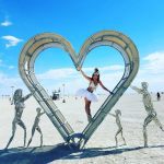 At Burning Man 2024, a woman in a white tutu and headdress balances within a large heart-shaped metal sculpture featuring two adults and two children holding hands. This scene, like many photos impossibles, unfolds on the vast desert under a bright blue sky.