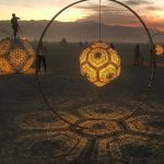 Large geometric lanterns with intricate patterns hang inside hoops, glowing and casting detailed shadows on the sand at dusk. Silhouettes interact with the lanterns, creating magical BurningMan photos against distant mountains and a colorful sky.