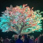 A large artificial tree with glowing leaves in vibrant pink, green, and blue stands against the night sky. Like a scene from Burning Man photos impossibles, crowds gather below, mesmerized by its brilliant lights and intricate branches.