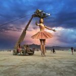 A giant ballerina marionette puppet in a pink tutu is suspended by cables from a crane on a wide desert plain at BurningMan 2024, with dramatic clouds, cyclists, and distant mountains—one of those photos impossibles.