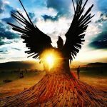 A dramatic wooden sculpture of a large winged figure stands outdoors at sunset, reminiscent of iconic BurningMan photos. The sun shines through the wings and scattered planks, while silhouetted people and bicycles dot the open landscape.
