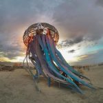 A large, artistic jellyfish sculpture with long, blue tentacles stands on a sandy desert at sunset. People and bicycles surround it as colorful lights glow—an iconic scene captured in BurningMan photos of surreal moments and photos impossibles.