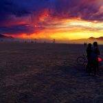 A dramatic sunset with vivid orange, yellow, and purple clouds lights up the vast desert at BurningMan. Silhouetted people and bicycles stand in the foreground, with others scattered beneath the colorful sky in these stunning BurningMan photos.