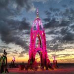 A tall, ornate art installation arches upward, glowing with pink and purple lights against a dramatic sunset sky at BurningMan. The sculpture stands on a sandy expanse, surrounded by clouds and smaller lit structures—true photos impossibles.