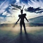 A silhouette of a person with twisted, horn-like headgear stands on a misty plain at sunset during BurningMan 2024. Dramatic clouds and sunlight stream behind them, capturing the mysterious energy of photos impossibles in this striking scene.