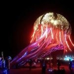 A giant, glowing jellyfish sculpture with a dome made of mirrored panels and long, illuminated tentacles stands on a dark open ground at night at BurningMan, surrounded by people and colorful lights—perfect for photos impossibles.