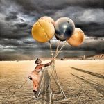 A person in festival attire and goggles stands on the sandy desert of Burning Man, holding large, colored balloons tied to poles. The cloudy sky is dramatic as balloon shadows stretch across the ground, with distant figures and structures beyond.