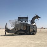 A large, metallic vehicle shaped like a dragon with an open mouth and spiked tail stands on a flat desert at BurningMan. Its body is made of panels and mesh, with a platform structure on its back. A person stands nearby for scale under the clear blue sky.