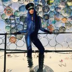 A person in a shiny blue bodysuit, tall platform boots, and a black hat with blue hair stands against a fence decorated with colorful plates at BurningMan. The background reveals a sandy landscape with scattered people and art installations.