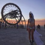 A woman in an ornate headpiece, bikini, and boots stands on a desert plain at sunset—a scene straight from BurningMan photos. Behind her, a massive metal wheel art structure teems with climbers as the sky glows orange and blue in these photos impossibles.