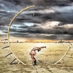 A person in festival attire bends backward gracefully under a large, crescent moon sculpture in the desert at Burning Man. The dramatic sky and distant art installations on the sandy playa create a scene worthy of photos impossibles.