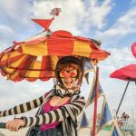 A smiling woman rides a bike with a colorful, striped umbrella hat at Burning Man. She wears large, flower-shaped sunglasses, a black-and-white scarf, and a pink top. In the background, there’s a giant pink flamingo sculpture and partly cloudy sky.