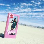 A person stands inside a large pink box resembling a Barbie doll package with BRC Edition and Barbie written on it, channeling classic BurningMan vibes. Sunglasses and a hat complete their look in this iconic BurningMan photos scene under the vast desert sky.
