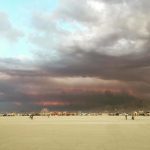 A wide, sandy desert with scattered people and vehicles in the distance under a dramatic sky. Thick, dark clouds with hints of red and purple loom overhead. Art structures evoke a festival, reminiscent of surreal BurningMan photos impossibles.