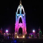 A tall, ornate archway glows with multicolored lights—pink, blue, and purple—against a dark night sky at BurningMan. Silhouetted people and bicycles gather around this striking structure, perfect for unforgettable BurningMan photos impossibles.