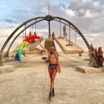 A woman in colorful festival attire and fishnet stockings poses in front of a large art swing at BurningMan. The sky is cloudy, people interact with the structure, and desert sand covers the ground—capturing true BurningMan photos energy.