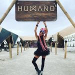 A woman with pink braids, goggles, black top, tutu skirt, and knee-high socks poses under a large wooden HUMANO THE TRIBE sign at an outdoor festival. This striking scene captures the photos impossibles spirit of Burning Man photos.