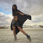 A person in a black hooded cape, chain top, studded skirt, and boots stands smiling on a sandy open area at Burning Man. They wear bracelets, a belt bag, and have thigh tattoos. Tents and domes dot the background beneath a cloudy sky.