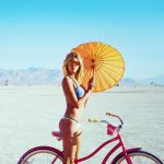 A woman in a blue bikini stands beside a pink bicycle on a flat, sandy landscape at Burning Man. She holds an orange parasol and wears sunglasses, with mountains and a blue sky in the background—a scene straight from photos impossibles.