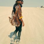 A person with long hair, wearing a leopard-print shirt, black pants, grey boots, and a black headband, walks on a wide, sandy landscape under a hazy sky—capturing that relaxed BurningMan vibe found in iconic BurningMan Photos.