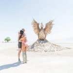 A couple embraces in the foreground on a sunlit, dusty plain at BurningMan. Behind them stands a large wooden phoenix sculpture with wings outstretched. A solitary tree and distant figures dot the pale desert landscape—true BurningMan Photos.