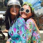 Two women smile and pose closely together at the BurningMan festival, wearing colorful, patterned bodysuits with My Little Pony and psychedelic designs. Both sport elaborate hats and sunglasses, including floral frames and a shiny sequined cap.