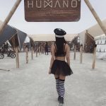 A woman in a black crop top, short skirt, police hat, and striped socks walks on dusty ground toward a “HUMANO THE TRIBE” sign at BurningMan, with tents, bikes, people, and art installations in the background.