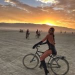 A woman in a black outfit, gloves, and boots sits on a bicycle on the flat desert at Burning Man sunset. Other cyclists trail behind as mountains are silhouetted against a dramatic sky—one of those photos impossibles to capture elsewhere.