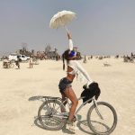 A woman poses with one leg up on a bicycle in the desert at Burning Man, holding a white parasol overhead. She wears a white top, blue shorts, boots, and blue goggles. Bikes, people, and art installations appear under a hazy sky—one of those photos impossibles.