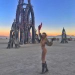 A woman in a sheer, sparkly outfit and boots stands on a desert playa at sunset, raising one arm and holding a red fan. Behind her are tall, ornate arch sculptures with flowers—an iconic scene from BurningMan photos.