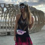 A woman stands outdoors at BurningMan, wearing a white fluffy cat ear headband, futuristic sunglasses, a black fishnet dress with pink fringe, and a shiny holographic fanny pack. Sculptural wooden art structures rise behind her under a partly cloudy sky.