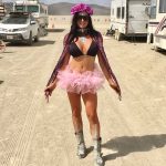 A woman stands on a dusty road between RVs at BurningMan, wearing a pink tutu, black bikini top, metallic boots, purple sunglasses, and a flower crown. She smiles with arms outstretched; sky and distant mountains frame this unique BurningMan photo.