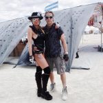 A woman and man stand arm in arm on sandy ground at an outdoor event, capturing the spirit of BurningMan. She wears a black hat, sunglasses, bodysuit, gloves, and boots; he sports a gray shirt and shorts. Tents and flags fill the background—true Burning Man photos.