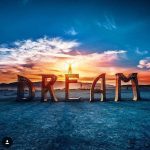 A person stands with arms raised atop large, illuminated letters spelling “DREAM” on a flat, sandy landscape at sunset—a magical scene that captures the surreal spirit found in BurningMan photos. Vibrant clouds and glowing mountains frame the dazzling sky.