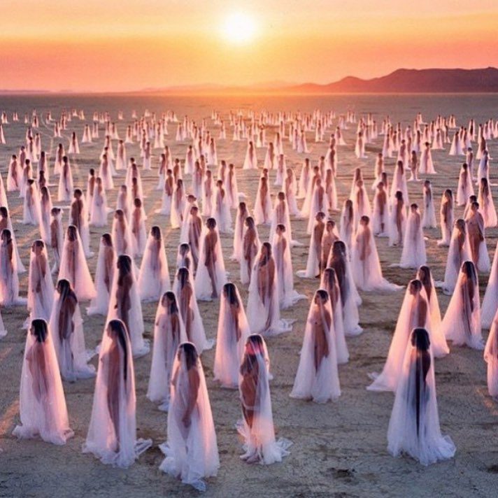 Hundreds of women draped in sheer white veils stand spaced apart on a sandy, barren landscape at sunset. Reminiscent of BurningMan photos, the surreal scene glows with soft pink and gold light beneath an orange sky and distant mountains.