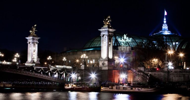 Night view of Paris’ Pont Alexandre III bridge with ornate lampposts and golden statues. Behind, the illuminated Grand Palais glows under the French flag—perfect sights to enjoy after settling into your accommodation or Hotel Beaubourg in Paris.