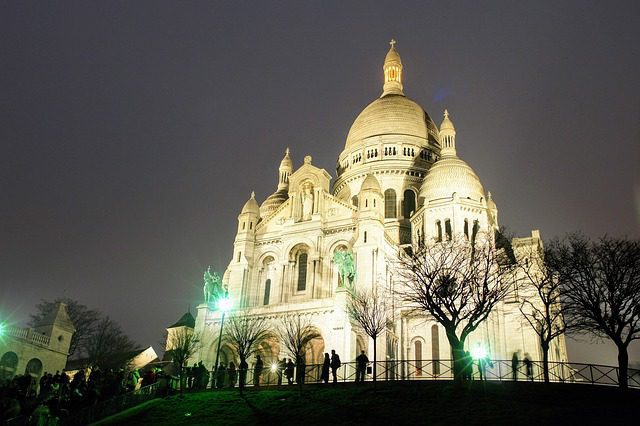 The illuminated Sacré-Cœur Basilica stands against a night sky, its white domes and ornate facade glowing. Silhouetted trees and a crowd of people are visible in the foreground on a grassy hill, with green lights casting accents on the scene.