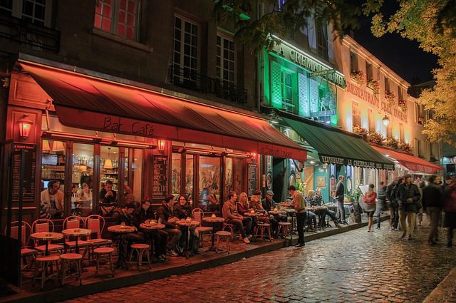 A lively Parisian street café at night with red and green awnings, glowing lights, and people sitting at outdoor tables on cobblestone pavement, chatting and dining. Others walk by under trees and the warmly lit restaurant windows.