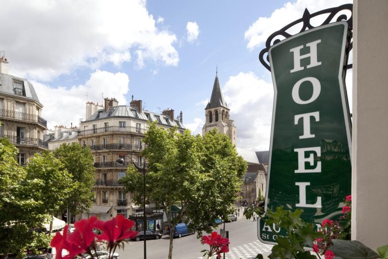 A large green Hôtel Au Manoir sign hangs on a building, overlooking a Parisian street in Saint-Germain lined with trees, historic stone buildings, and a church steeple. Red flowers in the foreground add color under a partly cloudy sky.