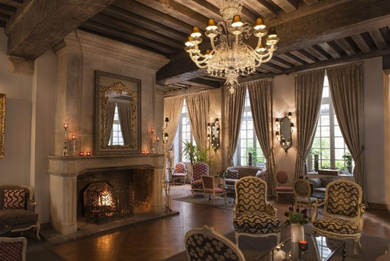 Elegant sitting room at Hotel d'Aubusson with ornate chairs, a lit stone fireplace, and a gilded mirror. A crystal chandelier hangs from wooden beams, while tall draped windows fill this Paris luxury hotel space with natural light.