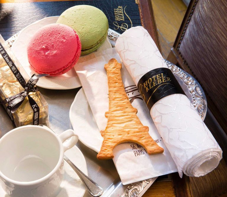A table setting at Hôtel Kléber features a white napkin wrapped in a black and gold ring, pastel macarons, a gold-wrapped chocolate, an Eiffel Tower cookie, and sunlight streaming in—reflecting the elegance of Paris hotel luxury accommodation.