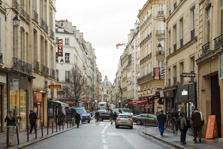 A lively Paris street scene in Saint-Germain, with cars and people along tall beige buildings featuring balconies, cafés, shops, and a Paris hotel. Signs for Hôtel Left Bank and a domed historic building rise above the bustling street.