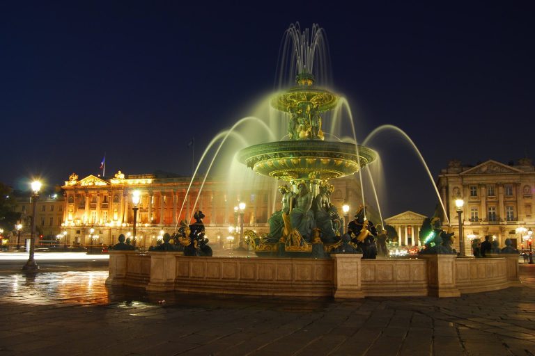 At night, a large ornate fountain with statues and multiple water streams is lit up in a grand city square near Hôtel Queen Mary. Elegant classical buildings glow under a deep blue sky, with street lamps and reflections shimmering on the wet pavement.