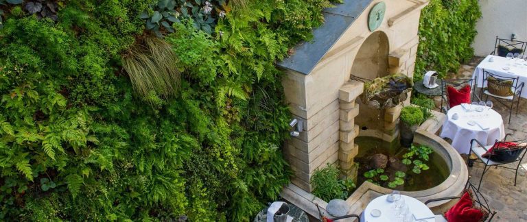 A lush vertical garden covers a wall beside outdoor dining tables. A stone fountain with water lilies and a small statue is framed by greenery. White tablecloths, glassware, and chairs with red and black cushions create an inviting, elegant patio atmosphere.