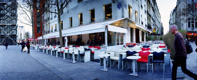 A sidewalk café with rows of empty tables and chairs in red, white, and blue sits beside a modern building. A few people walk by, bare trees line the street, and the urban scene evokes Paris—a spot any art lover’s guide would recommend.