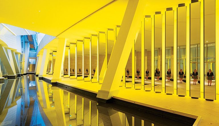 A modern hallway at the Fondation Louis Vuitton features tall, vertical yellow-lit columns reflected in a still water pool. Multiple women in dark outfits sit on benches, evoking a striking art installation of repetition and light.