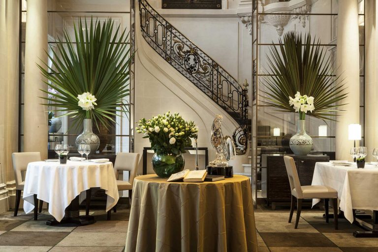 Elegant restaurant interior at Taillevent with white tablecloths, beige chairs, and a decorative round table in the center holding a lush bouquet of white roses—perfect for un anniversaire de 60 ans. Tall green palm leaves grace vases by the grand staircase.