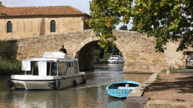 A small white houseboat and a blue rowboat float on a narrow canal beside an old stone arch bridge in France. A person stands on the houseboat as sunlight shines, with green trees and a rustic stone building in the background.